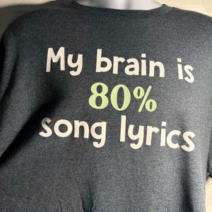May include: Dark gray t-shirt with the text "My brain is 80% song lyrics" in white and green. The shirt is displayed on a mannequin. The text is centered on the front of the shirt.