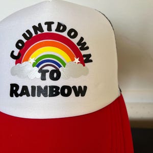 Peut inclure: Casquette de camionneur blanche et rouge avec un arc-en-ciel et le texte "Countdown to Rainbow".