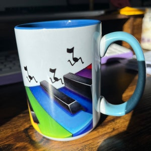 K&ouml;nnte beinhalten: Eine wei&szlig;e Keramiktasse mit blauem Rand und Henkel. Die Tasse zeigt ein farbenfrohes Klaviertasten-Design mit Noten, die &uuml;ber die Tasten laufen.