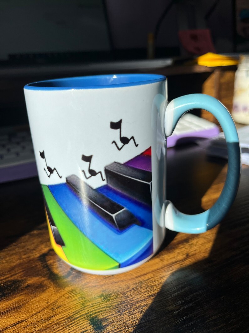 K&ouml;nnte beinhalten: Eine wei&szlig;e Keramiktasse mit blauem Rand und Henkel. Die Tasse zeigt ein farbenfrohes Klaviertasten-Design mit Noten, die &uuml;ber die Tasten laufen.