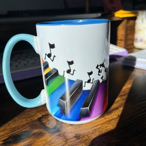 K&ouml;nnte beinhalten: Eine wei&szlig;e Keramiktasse mit blauem Rand und Henkel. Die Tasse zeigt ein farbenfrohes Design mit Klaviertasten und Noten.