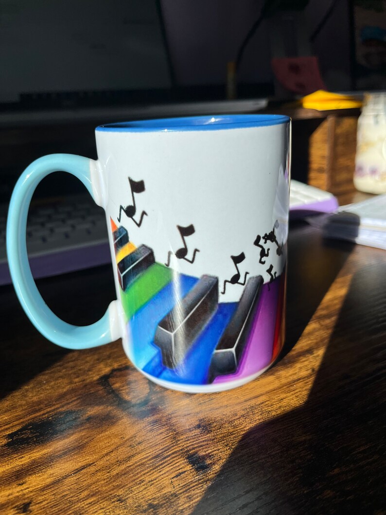 K&ouml;nnte beinhalten: Eine wei&szlig;e Keramiktasse mit blauem Rand und Henkel. Die Tasse zeigt ein farbenfrohes Design mit Klaviertasten und Noten.