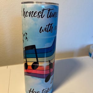 Puede incluir: Un vaso azul y blanco con un diseño de arcoíris y notas musicales. El texto en el vaso dice "An honest tune with a... Has taken".