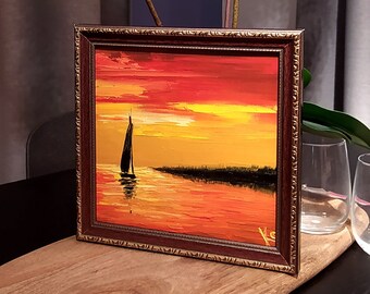 Può includere: Un dipinto incorniciato di una barca a vela su un mare al tramonto. Il dipinto è in una cornice marrone con una bordura dorata. Il dipinto è su un tavolo di legno con un vaso di orchidee bianche sullo sfondo.