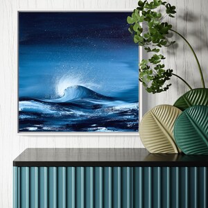 May include: A framed abstract painting of a blue wave crashing in the ocean. The wave is white at the crest and the background is a dark blue with white speckles. The painting is hanging on a white wall above a teal cabinet with black trim.