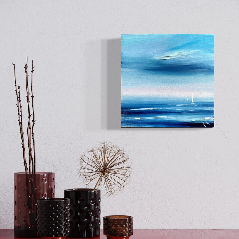 Op de afbeelding: Een schilderij van een zeegezicht met een zeilboot in de verte. De lucht is lichtblauw met witte wolken en het water is diepblauw. Het schilderij is ingelijst en hangt aan een witte muur.