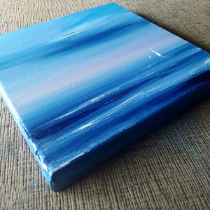 Op de afbeelding: Een klein, blauw canvas schilderij dat een zeezicht met een zeilboot in de verte afbeeldt. Het schilderij heeft een textuur, penseelstreek effect.