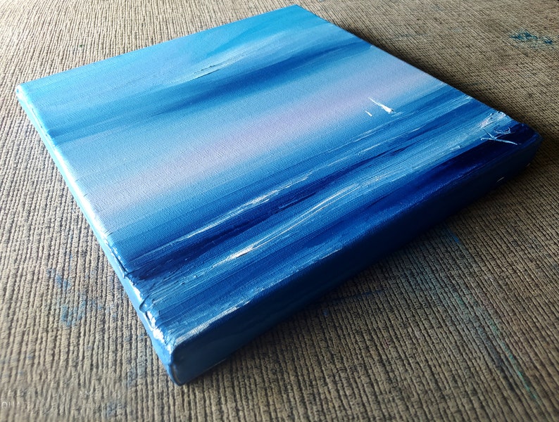 Op de afbeelding: Een klein, blauw canvas schilderij dat een zeezicht met een zeilboot in de verte afbeeldt. Het schilderij heeft een textuur, penseelstreek effect.