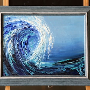 May include: A framed painting of a large blue wave crashing in the ocean. The wave is painted with thick, textured brushstrokes, giving it a dynamic and realistic look. The painting is framed in a light blue wood frame.