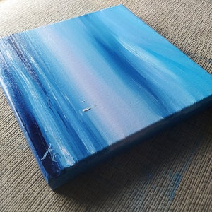 Op de afbeelding: Een klein, blauw en wit abstract schilderij op canvas. Het schilderij heeft horizontale penseelstreken die een gevoel van beweging en diepte cre&euml;ren.