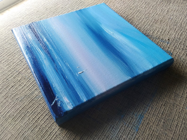 Op de afbeelding: Een klein, blauw en wit abstract schilderij op canvas. Het schilderij heeft horizontale penseelstreken die een gevoel van beweging en diepte cre&euml;ren.