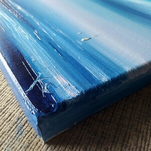 Op de afbeelding: Close-up van de zijkant van een blauw en wit abstract schilderij op canvas. Het schilderij heeft een textuur en is geschilderd met dikke penseelstreken.