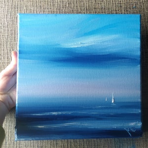 Op de afbeelding: Een schilderij van een zeezicht met een blauwe lucht en een lichtblauwe zee. Twee witte zeilboten zijn in de verte te zien.
