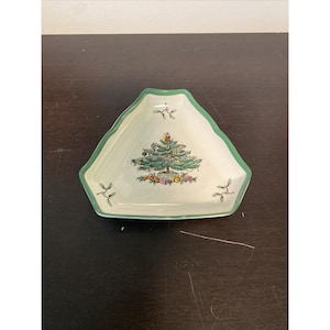 May include: A triangular, off-white ceramic dish with a green scalloped edge. The dish features a Christmas tree design with ornaments and wrapped gifts. Small green sprigs are placed around the rim. A festive holiday serving piece.
