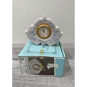 May include: A white porcelain clock with a gold-tone clock face and Roman numerals. The clock has a decorative floral design in light blue and is displayed on a blue box. The clock is approximately 4 inches wide.