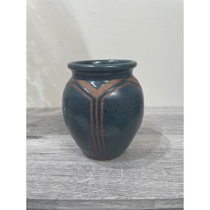 May include: A small, blue ceramic vase with a rounded body and a slightly flared rim. The vase features a brown geometric design around the upper portion. The vase is approximately 5 inches tall and is suitable for small floral arrangements.