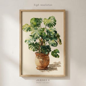 Puede incluir: Impresión en acuarela de una planta de Monstera en una maceta de terracota, sobre un fondo neutro. La obra de arte está enmarcada en un marco de madera clara. La impresión está etiquetada como "alta resolución" y "JERDELA PRINT STUDIO".