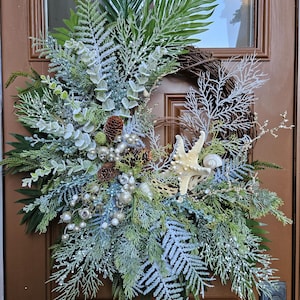 May include: A decorative wreath featuring a mix of green and frosted fern leaves, pinecones, silver ornaments, and seashells. A starfish and a seashell are prominent. The wreath is displayed on a brown door.