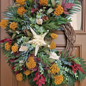 May include: A decorative wreath featuring a natural grapevine base adorned with artificial greenery, deep red foliage, and mustard yellow flower clusters. A large, textured cream-colored starfish and several small seashells are nestled among the plants.