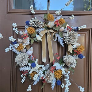 May include: A decorative wreath featuring white berries, dried flowers in mustard yellow and red, blue thistle, various seashells, and a white starfish. A central beige velvet ribbon hangs down, with the entire arrangement mounted on a brown door.
