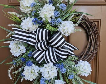 May include: A decorative wreath featuring white hydrangea blooms, blue accent flowers, and green foliage. A large black and white striped bow is centered on the wreath, which is made of woven brown branches. This floral wreath is perfect for a front door.