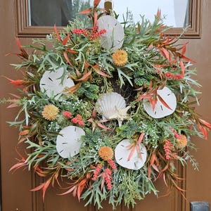 May include: A coastal-themed wreath featuring white sand dollars, seashells, and a starfish. The wreath is adorned with green and orange foliage, and small orange and yellow flowers. It is hanging on a brown door.