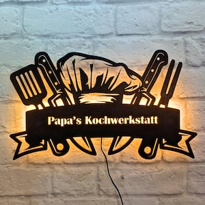 May include: A black metal sign with a chef's hat and kitchen utensils, illuminated from behind. The sign reads "Papa's Kochwerkstatt".