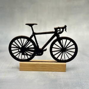 May include: A black bicycle silhouette, mounted on a rectangular wooden base. The bicycle features a classic road bike design with drop handlebars and spoked wheels. The wheels, seat, and frame are all black.
