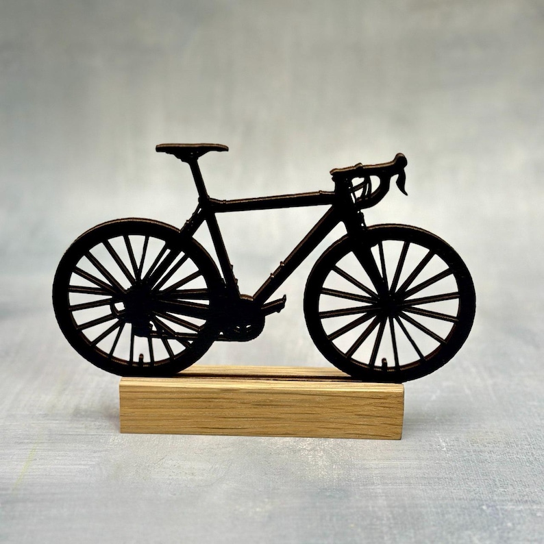 May include: A black bicycle silhouette, mounted on a rectangular wooden base. The bicycle features a classic road bike design with drop handlebars and spoked wheels. The wheels, seat, and frame are all black.