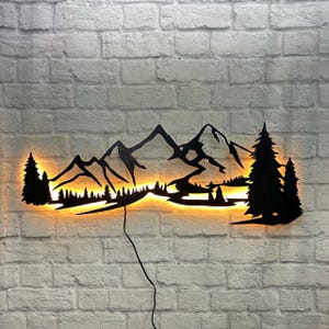 Peut inclure: Décoration murale en métal noir représentant une chaîne de montagnes avec un lac et des arbres. L'œuvre est éclairée par des lumières LED blanc chaud.