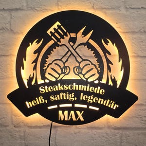 Könnte beinhalten: Schwarzes Metallschild mit einer stilisierten Darstellung von gekreuzten Grillwerkzeugen und Flammen. Der Text "Steaksmiede heiß, saftig, legendär MAX" ist auf dem Schild geschrieben.