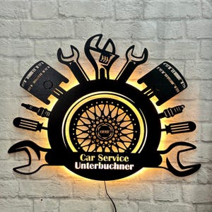Peut inclure: Art mural en métal noir représentant une roue de voiture, des clés et des pistons. L'œuvre est éclairée d'une lumière jaune chaude et porte le texte "Car Service Unterbuchner" sous la roue.