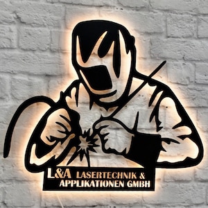 Op de afbeelding: Zwarte metalen silhouet van een lasser met een lasbrander, verlicht van achteren. De silhouet is gemonteerd op een witte bakstenen muur met een bord waarop "L&A Lasertechnik & Applikationen GmbH" staat.