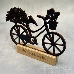 May include: A decorative wooden bicycle silhouette with a bouquet of flowers and a bird on the back. The bicycle has a basket with heart-shaped decorations and is mounted on a wooden base. The base has the text "Fahr Rad, sei frei!"