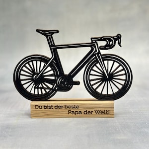 May include: A black bicycle-shaped wooden decoration. The bicycle has detailed wheels, a seat, and handlebars. It is mounted on a rectangular wooden base with the text "Du bist der beste Papa der Welt!"