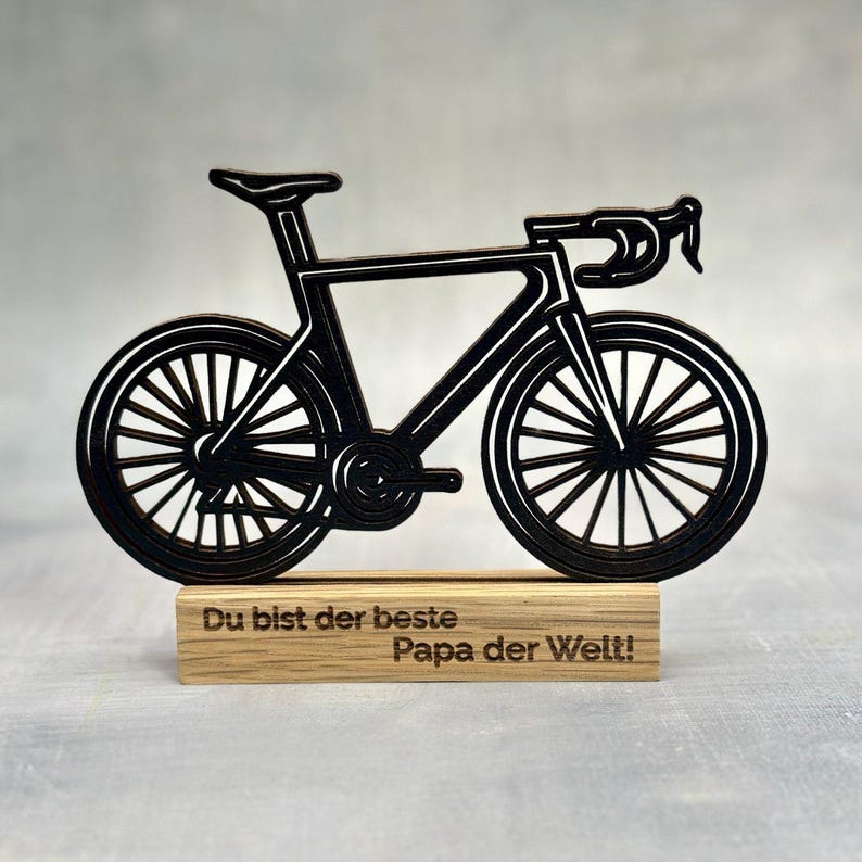 May include: A black bicycle-shaped wooden decoration. The bicycle has detailed wheels, a seat, and handlebars. It is mounted on a rectangular wooden base with the text "Du bist der beste Papa der Welt!"