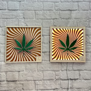 Op de afbeelding: Twee houten wanddecoraties met een groen marihuanabladontwerp. Het ontwerp is een draaiende patroon van bruine en witte strepen. Het linker stuk is een gewone wanddecoratie, terwijl het rechter stuk een verlichte wanddecoratie is. Beide stukken hebben de tekst "WP Wood Products" in de rechterbenedenhoek.
