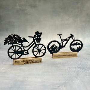 May include: Two black wooden bicycle decorations. One features a bicycle with flowers and a bird, the other a mountain bike with mountain silhouettes in the wheels. Each sits on a wooden base with German text.