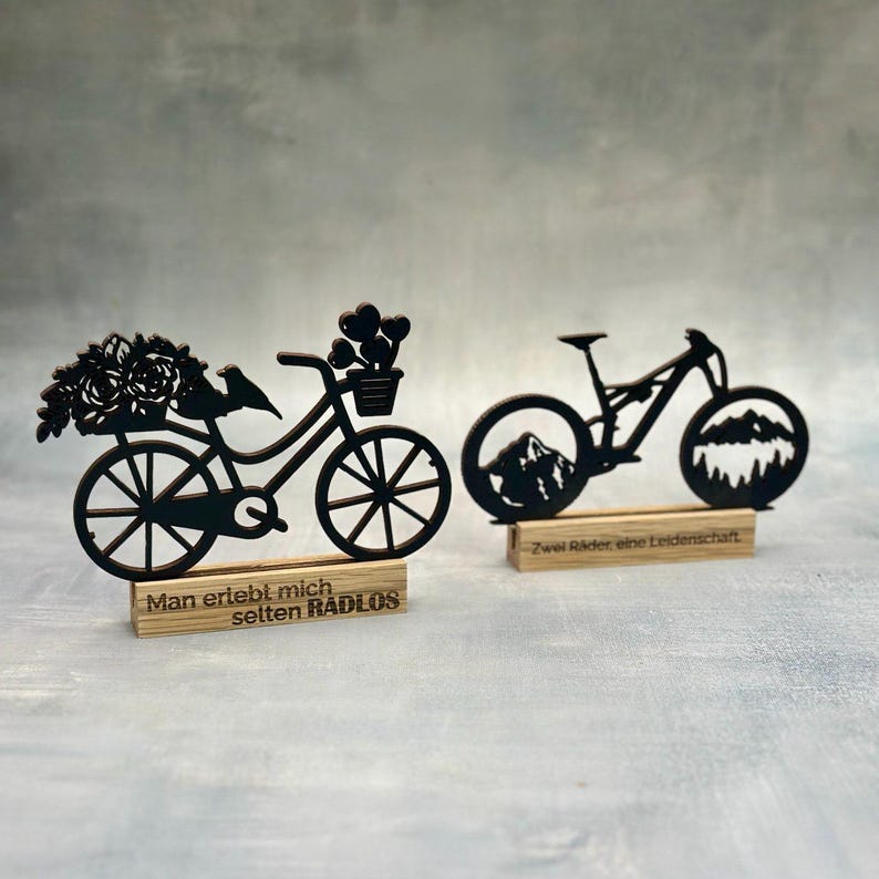May include: Two black wooden bicycle decorations. One features a bicycle with flowers and a bird, the other a mountain bike with mountain silhouettes in the wheels. Each sits on a wooden base with German text.