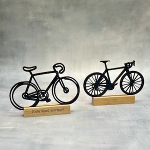 May include: Two black bicycle silhouettes on wooden bases. One has the text "Fahr Rad, sei frei!" on its base. The bicycles are detailed with handlebars, seats, and wheels. The background is a mottled gray.