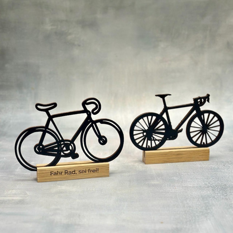 May include: Two black bicycle silhouettes on wooden bases. One has the text "Fahr Rad, sei frei!" on its base. The bicycles are detailed with handlebars, seats, and wheels. The background is a mottled gray.