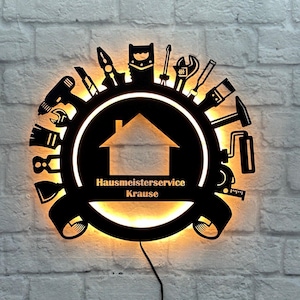 Könnte beinhalten: Schwarze Wandleuchte aus Metall mit einem runden Design, das verschiedene Werkzeuge und eine Haussilhouette zeigt. Die Leuchte ist mit einem warmen gelben Licht beleuchtet und trägt die Aufschrift "Hausmeisterservice Krause".