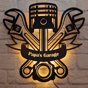 Könnte beinhalten: Schwarze Wandkunst aus Metall mit einem stilisierten Motor-Kolben mit Schraubenschlüsseln und einem Banner mit der Aufschrift "Papa's Garage".