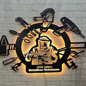 Pode incluir: Arte de parede de metal preto com um soldador com uma máscara de solda rodeado de ferramentas, incluindo um martelo, alicates, uma chave e uma broca. A arte é iluminada com uma luz quente e inclui o texto "250 Jahre Metallbau Weinmann".