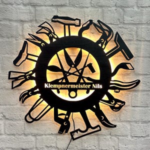 Könnte beinhalten: Schwarzes Metallschild mit einem kreisförmigen Design, das verschiedene Werkzeuge wie Hämmer, Zangen und Scheren zeigt, die um einen zentralen Kreis angeordnet sind. Der Text "Klempnermeister Nils" wird in der Mitte angezeigt und von einem warmen, gelben Licht beleuchtet.