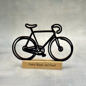 May include: A black bicycle silhouette cut from wood, mounted on a wooden base. The bicycle features a classic road bike design. The base has the text "Fahr Rad, sei frei!" in a dark font.
