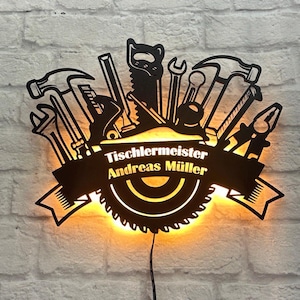 Pode incluir: Arte de parede de metal preto com uma lâmina de serra circular com uma faixa que diz "Tischlermeister Andreas Müller" e uma coleção de ferramentas que incluem um martelo, uma chave e uma serra. A arte é iluminada com luz amarela quente.