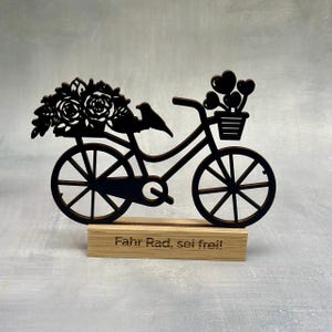 May include: A black wooden bicycle decoration with a basket of hearts and flowers. A small bird sits on the frame. The base is a wooden block with the words "Fahr Rad, sei frei!" engraved on it.