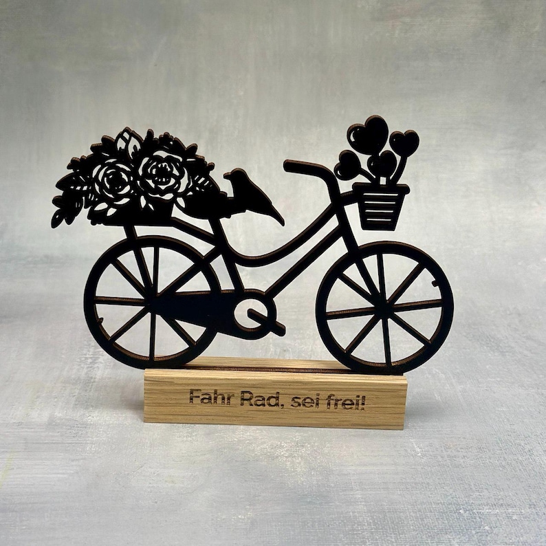 May include: A black wooden bicycle decoration with a basket of hearts and flowers. A small bird sits on the frame. The base is a wooden block with the words "Fahr Rad, sei frei!" engraved on it.