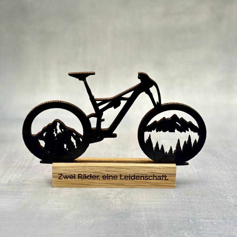 May include: A dark brown metal mountain bike sculpture with mountain and forest cutouts in the wheels, mounted on a wooden base. The base has the text "Zwei R&auml;der, eine Leidenschaft."